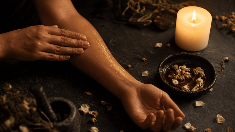A bare forearm resting on dark slate with the opposite hand gently rubbing warm amber oil into the skin beside a small ceramic dish of oil and dried petals, lit by a single candle — somatic self-massage for nervous system soothing.